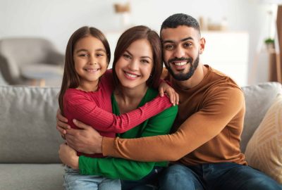 Portrait of cheerful Arabic family of three people hugging sitting on the couch at home, posing for photo and looking at camera. Smiling young bearded man embracing his loving wife and daughter