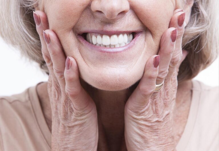 A woman smiles broadly while gently holding her cheeks with both hands, showing healthy, white teeth.